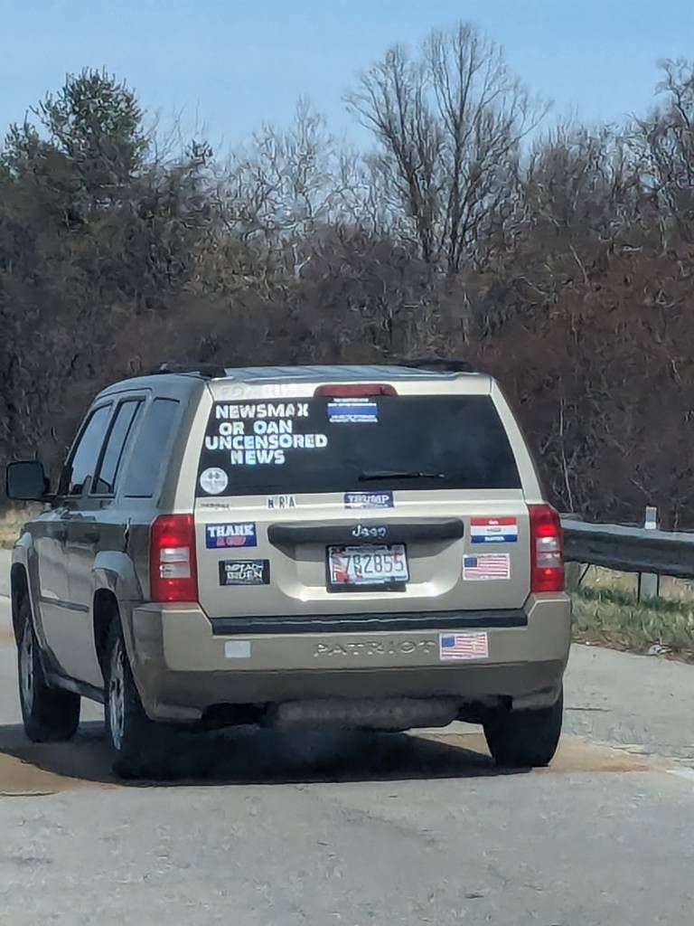 A tan Jeep Patriot with letters that spell out "NEWSMAX OR OAN UNCENSORED NEWS" A thin blue line sticker that is illegible, a "Thank God" Sticker, a Trump sticker, and some other things I can't make out. Would it be too malicious to add the license plate number here as well? 