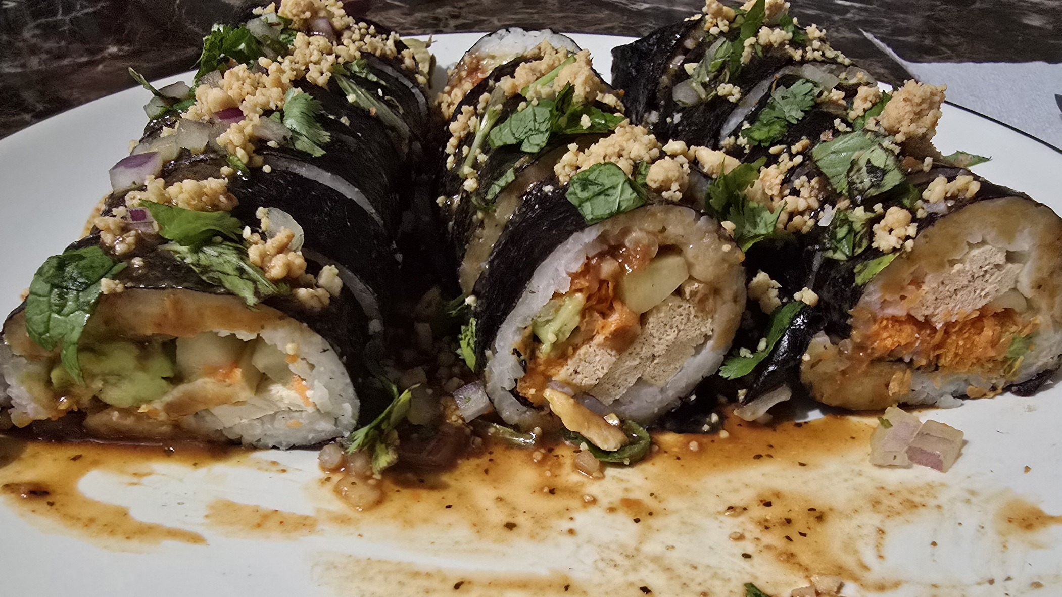 Three sushi rolls with rice, cucumber, carrot, cucumber, tofu, and Avocado. Topped with diced thai basil, cilantro, mint, and red onion, chopped peanuts, and a Thai peanut sauce.  