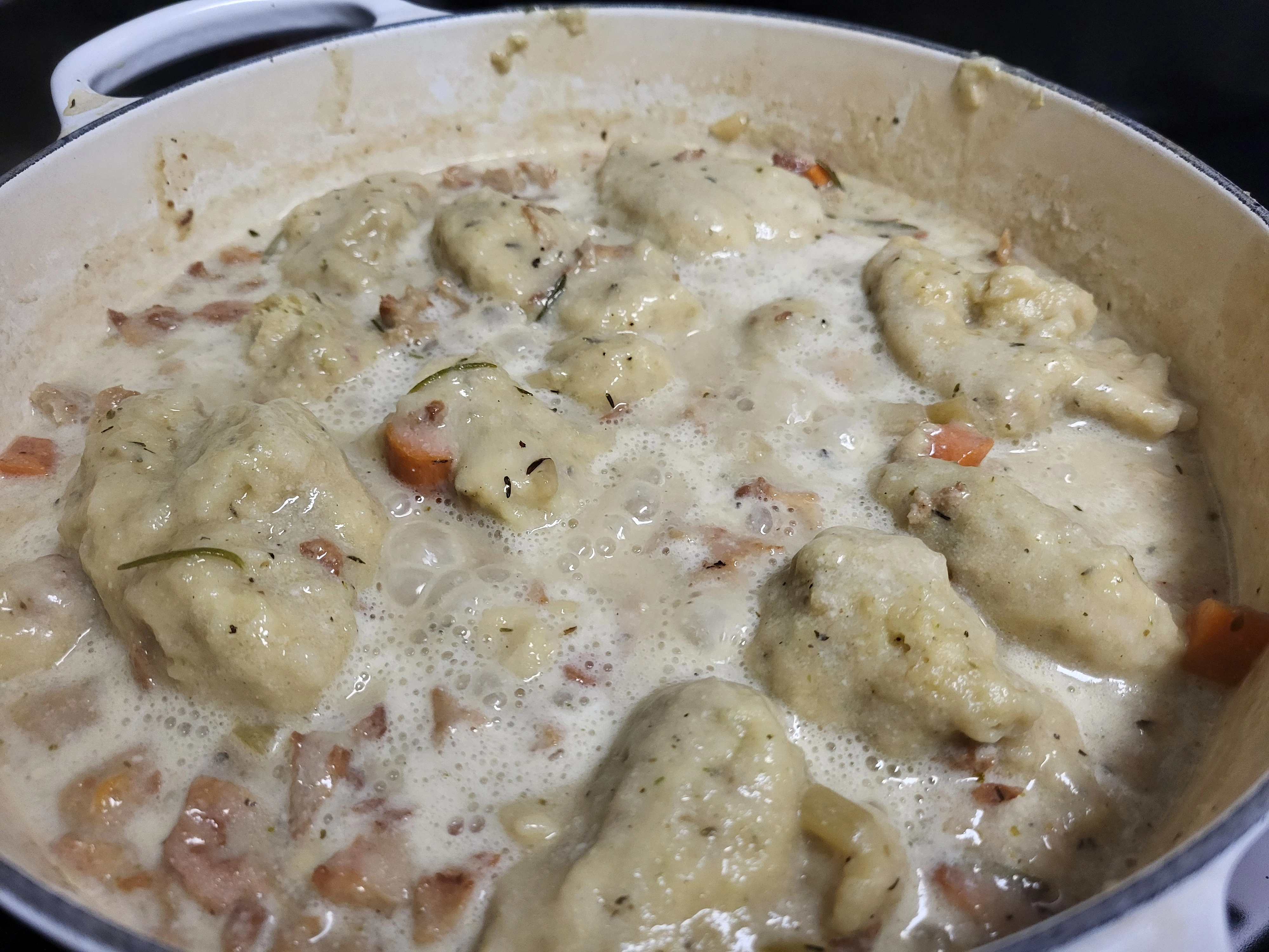 dutch oven full of a chicken and dumpling soup, little bits of carrots and seitan are visible as well as herbs, carrots, and large dumplings which have been boiled in the soup